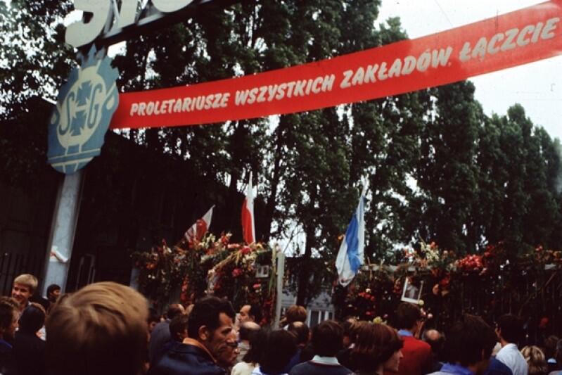 Władze PRL starały się doprowadzić do rozbicia solidarności między strajkującymi zakładami. W odpowiedzi liderzy MKS apelowali o to, by trzymać się razem. Hasło na transparencie nawiązuje do słynnego zdania z "Manifestu komunistycznego" Marksa i Engelsa: "Proletariusze wszystkich krajów łączcie się!". We wtorek, 19 sierpnia, do MKS należało już 200 zakładów