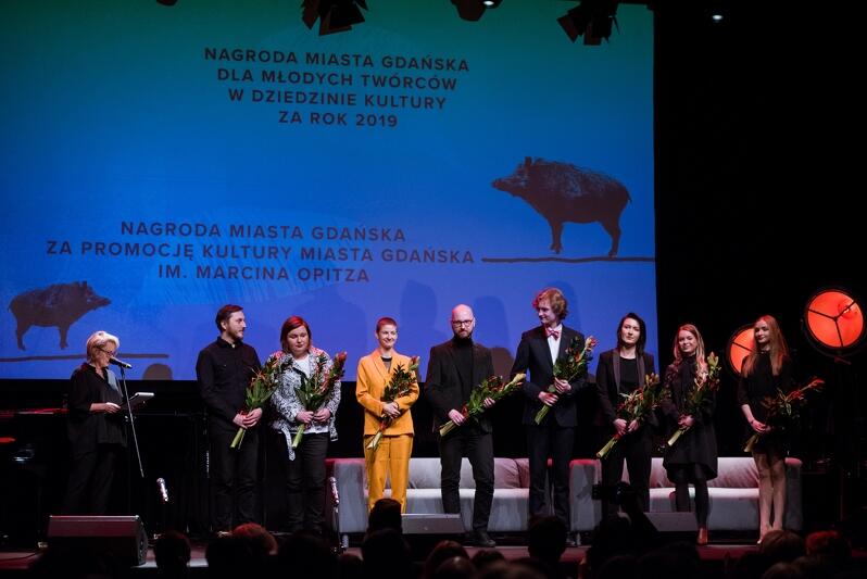 Na scenie, na tle wyświetlonego napisu Nagroda Miasta Gdańska dla Młodych twórców w dziedzinie kultury stoi osiem nominowanych osób, w rękach trzymają kwiaty. Po lewej stoi kobieta z krótkimi blond włosami przy mikrofonie.