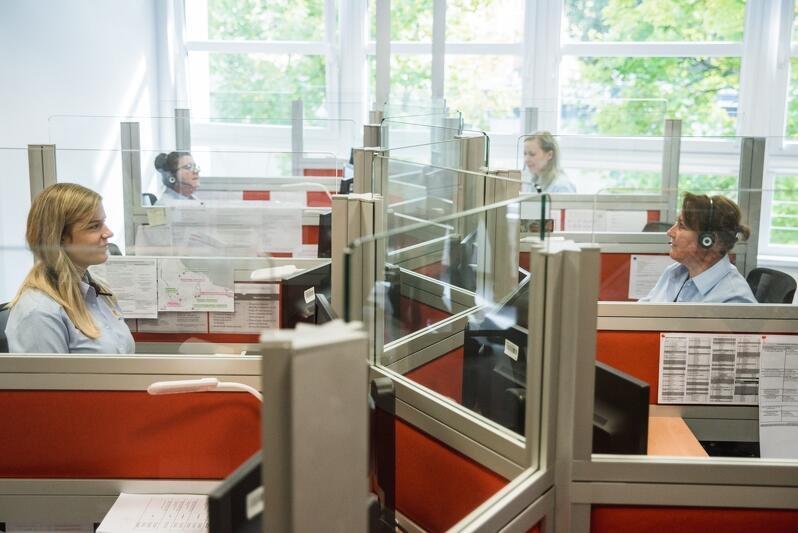 Wnętrze Gdańskiego Centrum Kontaktu, widać kilka osób siedzących przy komputerach, w słuchawkach z mikrofonami na uszach, oddzielają ich od siebie wzajemnie szyby z pleksi