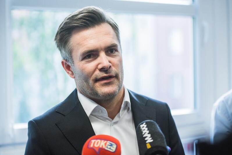 Siwiejący szatyn w średnim wieku w czarnej marynarce i białej koszuli wypowiada się podczas konferencji prasowej, przed nim widać dwa mikrofony dziennikarzy radiowych