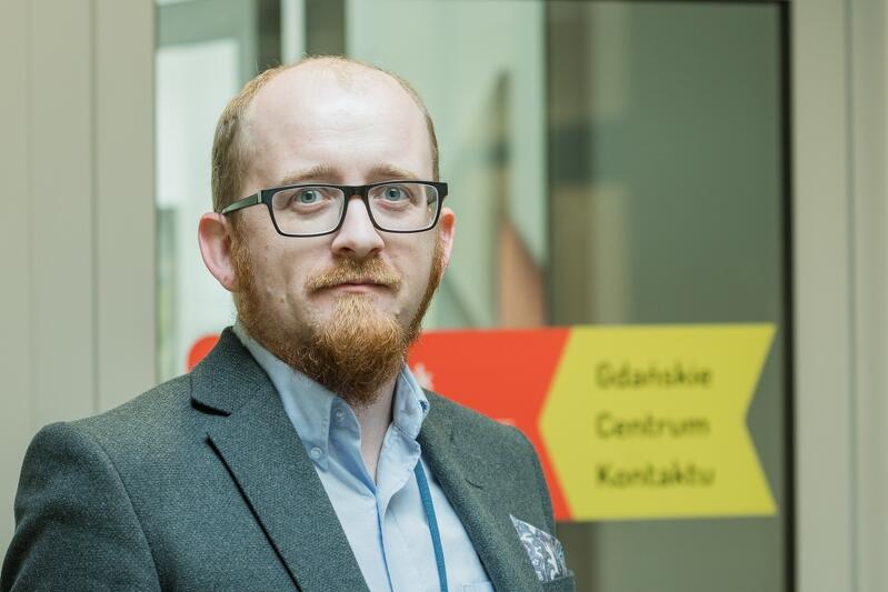 Mężczyzna w średnim wieku, rudy, mocno łysiejący, z brodą, w okularach, ubrany w szary garnitur wpatruje się w obiektyw
