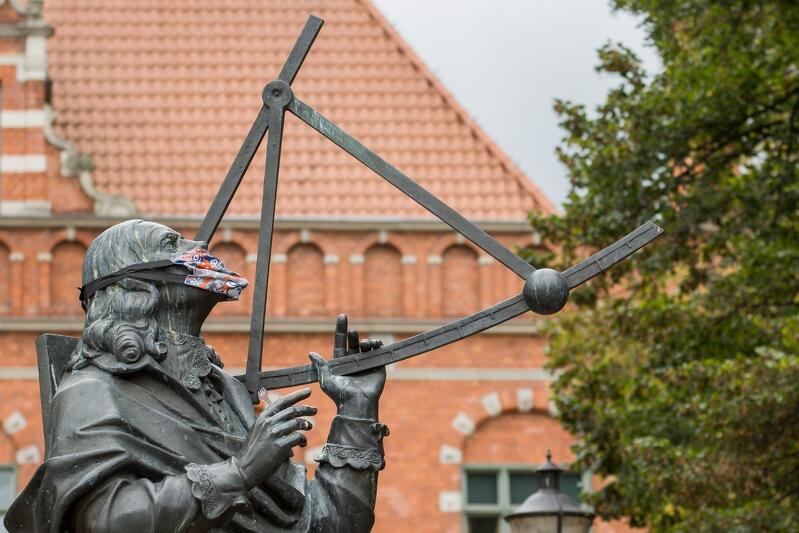 Twarz pomnika Heweliusza ubrana w maseczkę. W tle widoczny jest gmach Ratusza Staromiejskiego