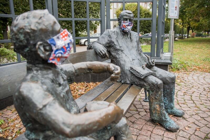 Obie postaci siedzą na dwóch skrajach pomnikowej ławeczki. Oskar na pierwszym planie - Grass wudoczny jest w głębi. Obaj są w kolorowych maseczkach