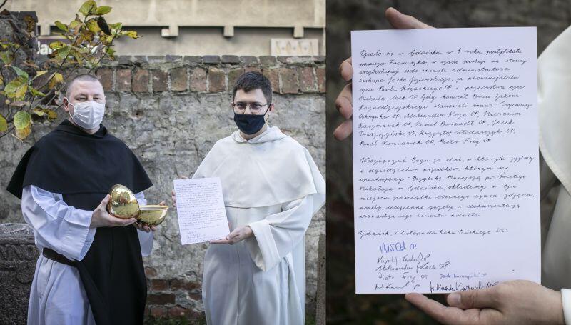 Dwaj ubrani na biało zakonnicy, jeden trzyma i prezentuje list, drugi otwartą kulę kapsuły