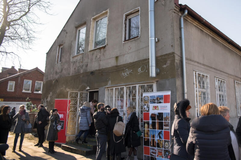 Na zdjęciu znajduje się budynek, pod którym stoi dużo osób ubranych w jesienne kurtki, zgromadzone osoby podzielone są na kilka grup. Rozmawiają ze sobą. Widać też, że jest przeprowadzany wywiad przed kamera. Przed wejściem do budynku stoi rollup reklamowy Gdańska