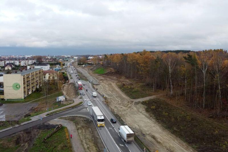 Jesień, drzewa bez liści, chmury, z lotu ptaka widać po lewej starą ulicę, którą jadą auta, a po prawej ślad nowej drogi, zerwana darń, góry piachu, powstająca warstwa podbudowy