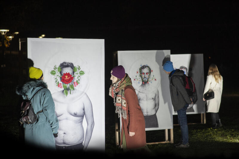 Wystawa plenerowa - na trzech dużych białych planszach widać grafiki przedstawiające sylwetki. Od lewej grafika mężczyzny z lekką nadwagą i czerwonym kwiatem w centralnej części twarzy, dookoła którego narysowane są zielone liście. Środkowa plansza przedstawia szczupłego mężczyznę z drobnymi kwiatami wokół twarzy. Obaj są rozebrani od pasa w górę. Trzecia grafika niewidoczna. Planszom przyglądają się przechodnie, widoczne są trzy kobiety w jesiennych płaszczach i mężczyzna w czarnej kurtce i niebieskiej czapce