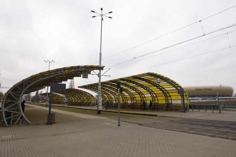 Tory tramwajowe, po bokach przystanki tramwajowe z duzymi żółtymi wiatami. W oddali fragment bursztynowego stadionu
