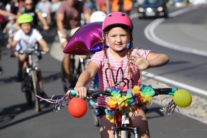 dziewczynka na rowerze, macha ręką do fotografa, różowy kask, warkoczyki, baloniki dla ozdoby na kierownicy i z tyłu roweru