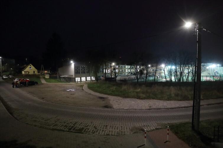 Na zdjęciu widać fragment drogi z nawierzchnią z płyt typu jomb, obok niej zarośnięty teren zielony, a w tle widać oświetlony budynek szkoły