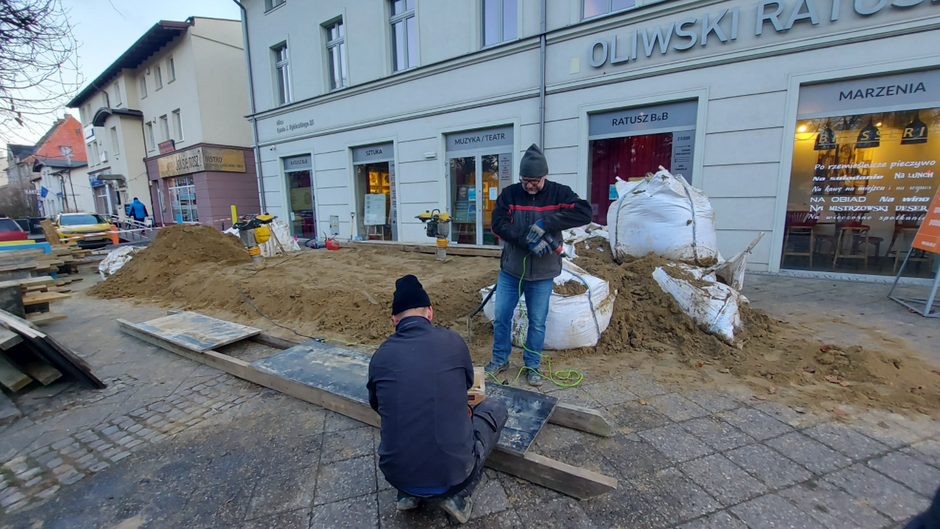 Na zdjęciu widać piasek rozsypany na chodniku i dwóch schylających się mężczyzn. W tle - jasna kamienica
