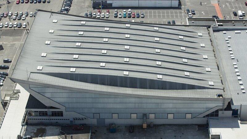 Zdjęcie z góry: dach i bok hali sportowej, obok po lewej oraz powyżej fragment parkingu z samochodami