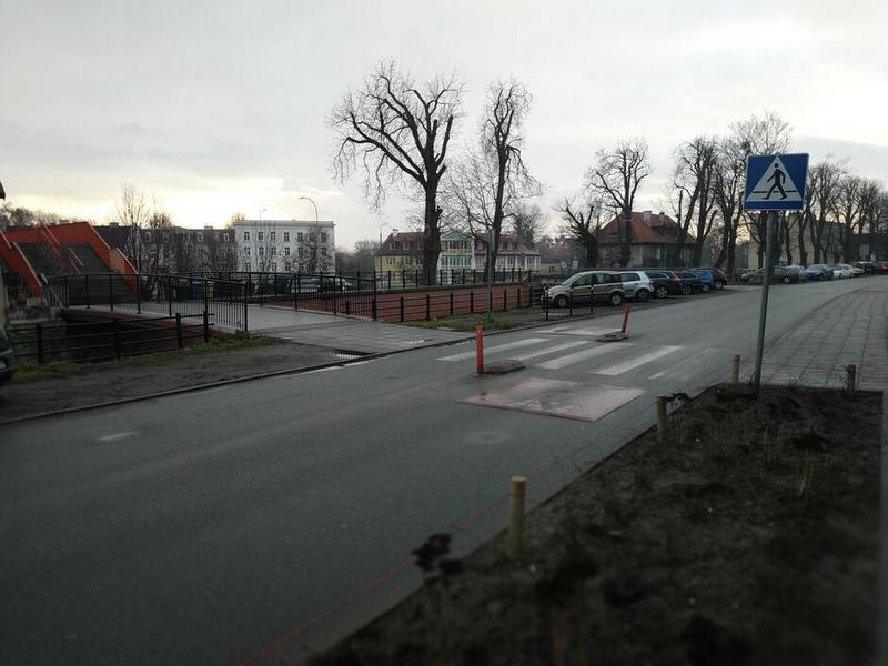 Ulica, na niej przejście dla pieszych. Przed przejściem - w obu kierunkach - progi zwalniające oraz słupki na środku jezdni. Po prawej stronie fragment pobocza i chodnika (przy przejściu dla pieszych), po lewej chodnik, pas parkingu z samochodami, dalej wiadukt dla pieszych i budynki