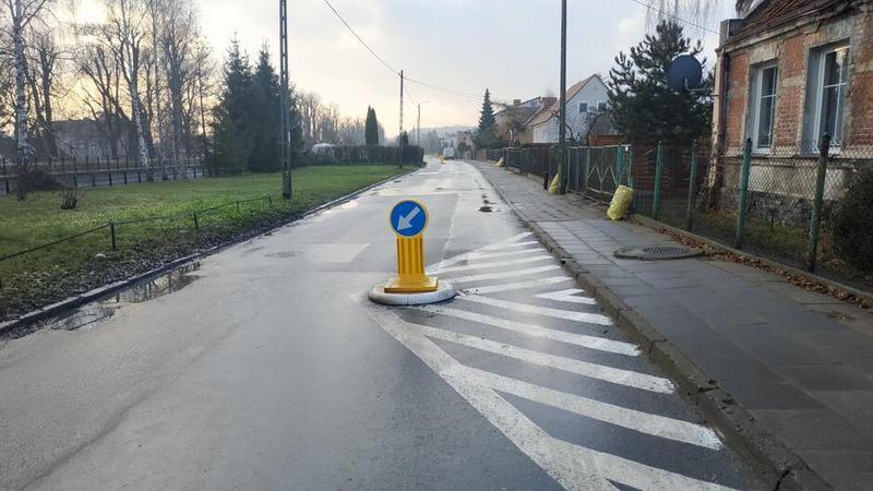 Ulica, obok po prawej chodnik, dalej płot i fragment domu z czerwonej cegły. Po lewej trawnik. Na ulicy wymalowany trójkąt oraz ustawiona szykana, zawężająca szerokość jezdni