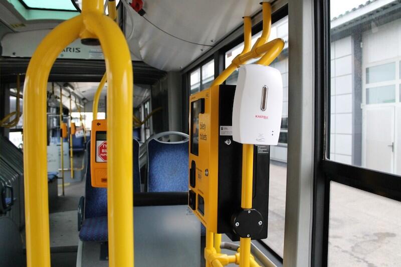 Wnętrze autobusu, widoczny biały dozownik, bordowe siedzenia i żółte poręcze