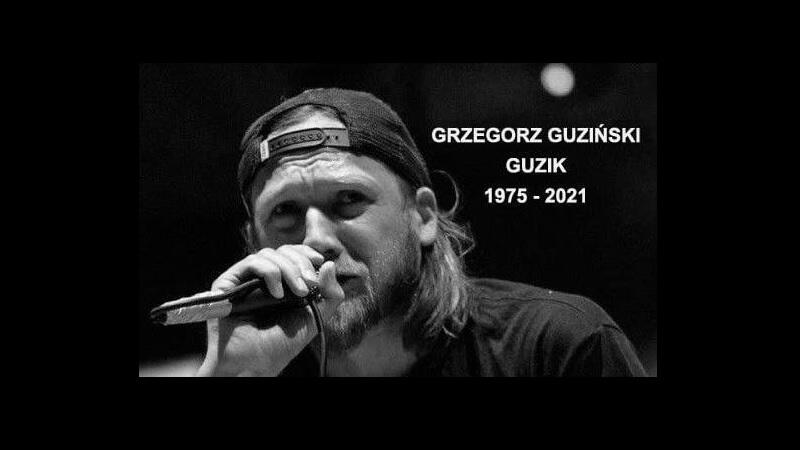 Twarz mężczyzny w średnim wieku w czapce z daszkiem (daszek odwrócony - niewidoczny), przed nim mikrofon, który trzyma dłonią. Mężczyzna śpiewa