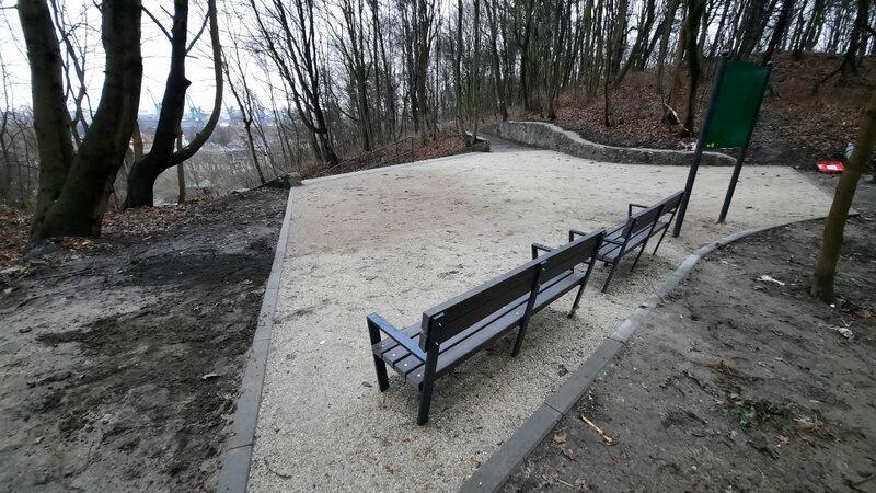 Na zdjęciu widać niewielki placyk o nawierzchni piaskowej, stoją tu dwie brązowe ławki