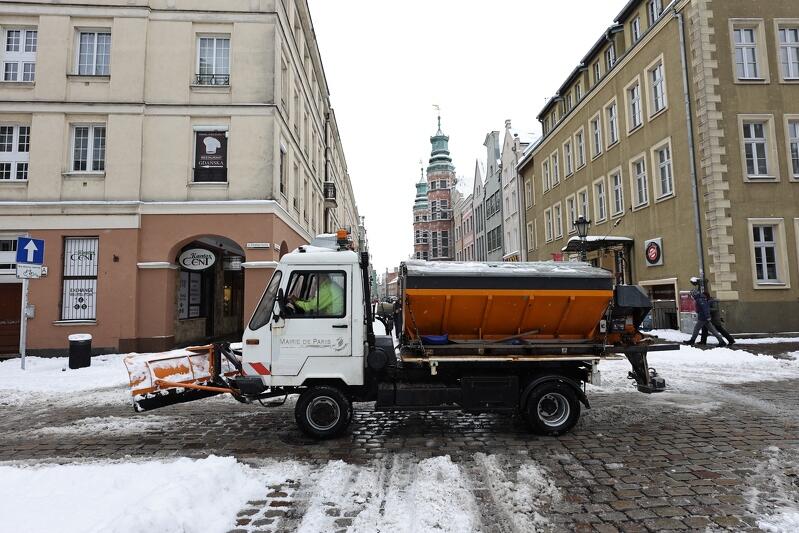 Pługopiaskarka w zabytkowym centrum Gdańska, dookoła zaśnieżone ulice chodniki i samochody