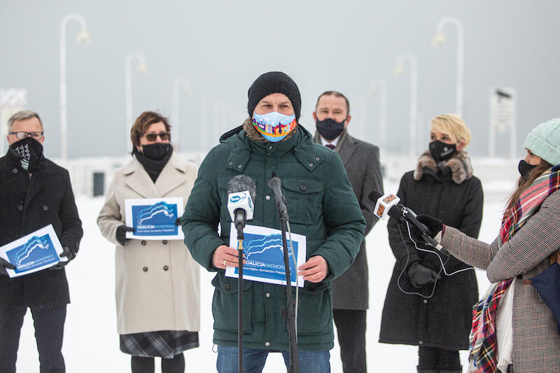 Mężczyzna w zielonej kurtce i kolorowej masce stoi przy mikrofonie