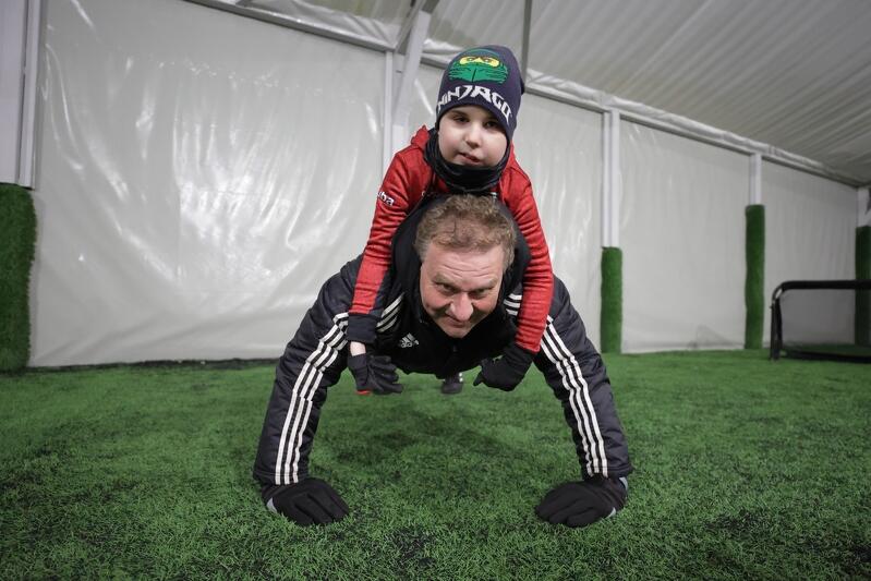 Dziecko leży na dorosłym, który w pozycji pompka jest na murawie. Oboje ubrani w zimowe stroje sportowe