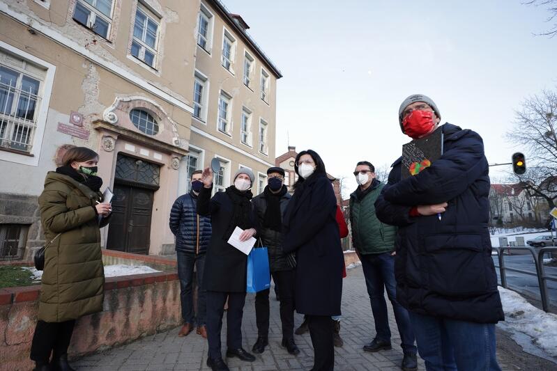 siedem osób w ubraniach zimowych i w maseczkach na twarzach patrzy gdzieś poza fotografa, po lewej stronie w tle budynek szkoły ze zniszczoną beżową elewacją