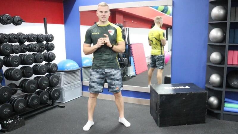Mężczyzna w stroju sportowym stoi w sali na siłowni. Za nim lustro, obok po obu stronach przyrządy do ćwiczeń: hantle, klocki, maty, skrzynia, kule