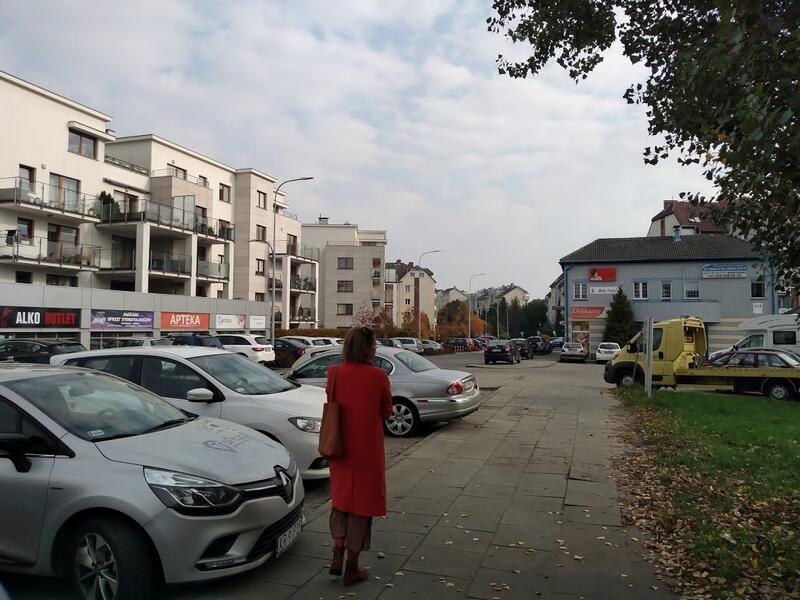 Chodnik, którym idzie kobieta, obok po lewej parking z samochodami. Za nim budynki mieszkalne 2-3 piętrowe, na wprost budynek jednopiętrowy