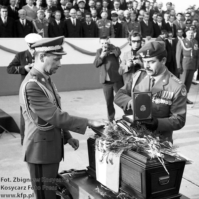 czarno-białe zdjęcie, dwóch wojskowych nad małą trumną, jeden z nich kładzie order na trumnie, w tle trybuna honorowa