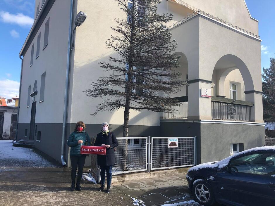 Na zdjęciu widać dwie kobiety w zimowych ubraniach, trzymają czerwoną tablicę z napisem rada dzielnicy, za nimi stoi jasna, duża kamienica