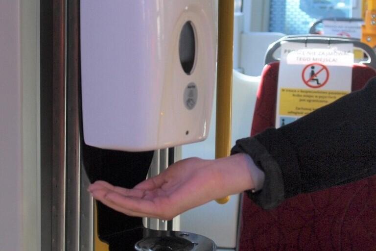 Wnętrze autobusu. Do ścianki przymocowany dozownik z płynem odkażającym, przy nim dłoń