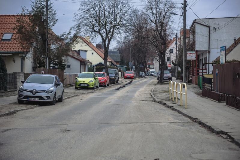 na zdjęciu fragment asfaltowej jezdni i chodnik, w tle zaparkowane samochody osobowe oraz niska zabudowa jednorodzinna