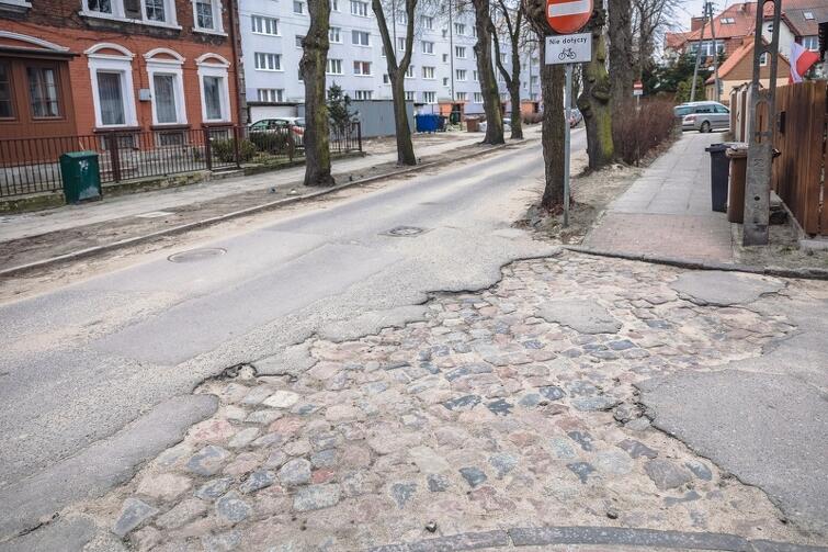 na zdjęciu fragment zniszczonej jezdni, w tym sporej wielkości wgłębienie z której wystają kamienie, w tle widać zaparkowane samochody i dom jednorodzinny oraz blok mieszkaniowy