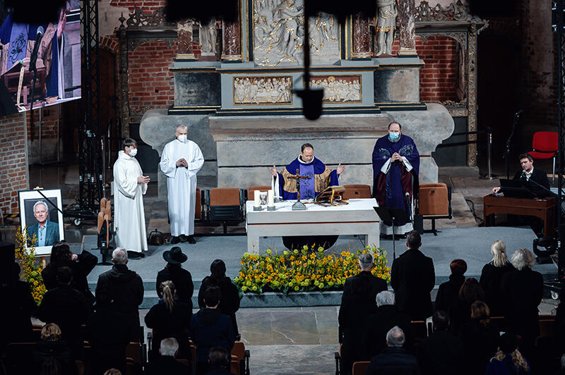 Wnętrze kościoła. Ksiądz w szatach liturgicznych stoi za stołem, ma rozłożone na boki ręce. Za nim inni księża oraz ołtarz. Przed ludzie w ławkach