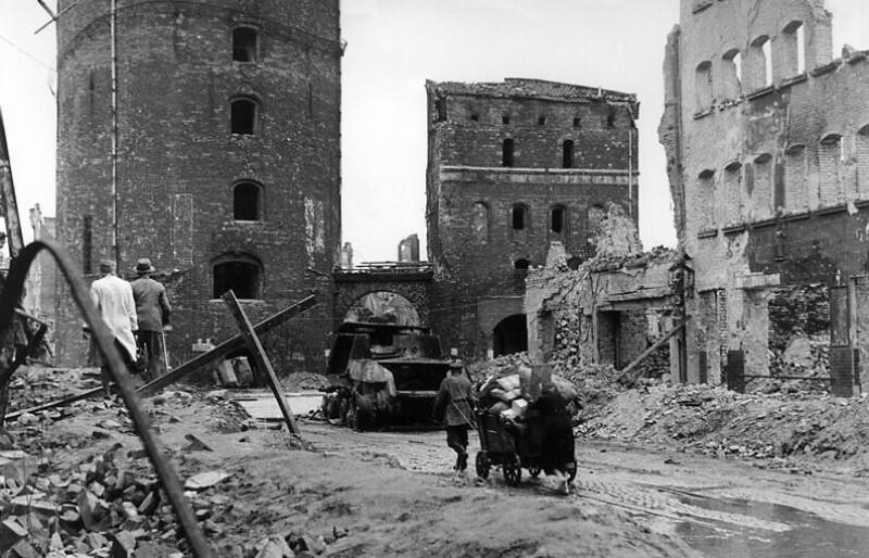 Wiosna 1945 roku. Środkiem ulicy Stągiewnej dwie osoby ciągną wózek, są obrócone plecami do widza. Po obu stronach ulicy widać zniszczone, częściowo zburzone budynki. Z lewej znajduje się sterta gruzu po której idą dwie inne osoby. W głębi ulicy widać wypalony transporter wojskowy. Ulicę zamyka sylwetka Bramy Stągiewnej