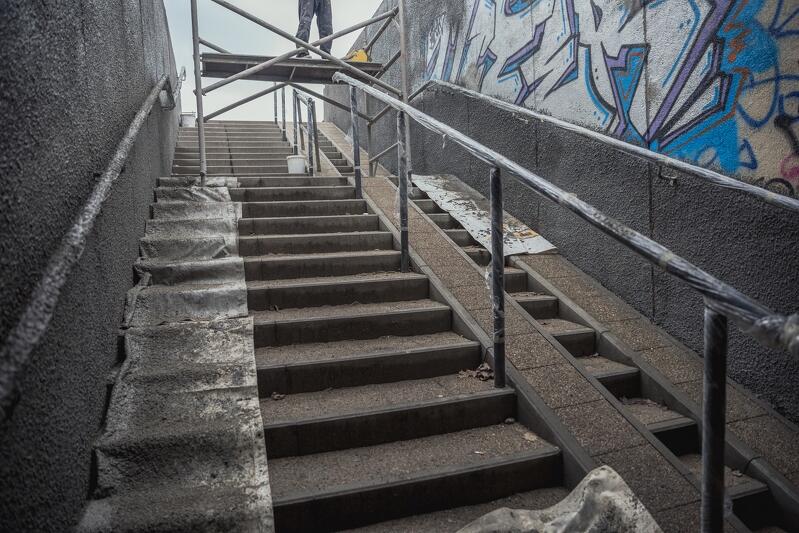Przejście podziemne widziane od środka. Schody prowadzone do góry, zabezpieczone taśmą poręcze i fragment schodów. Po środku rusztowanie, na nim widoczne nogi pracownika