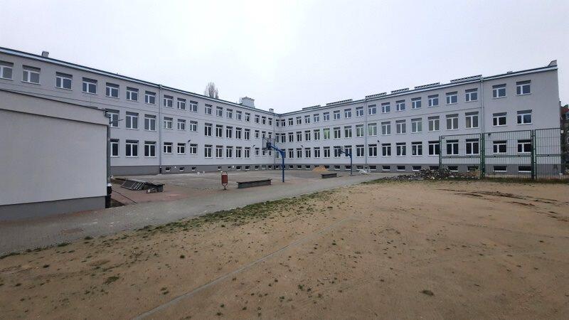 na zdjęciu jasnoszary budynek szkoły, trzypoziomowy w kształcie litery L, przed budynkiem widać fragment ubitego gruntu, jest też betonowy plac z zamontowanymi słupkami i koszami do gry w piłkę koszykową