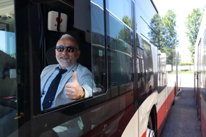 Prawy bok miejskiego autobusu, pokazany od strony kabiny kierowcy. Jest lato, upał. Przez uchylony boczny lufcik widać twarz uśmiechniętego kierowcy w okularach przeciwsłonecznych. Ubrany jest w niebieską koszulę, ma granatowy krawat. Pozuje do zdjęcia trzymając w okienku zaciśniętą dłoń z wystawionym do góry kciukiem