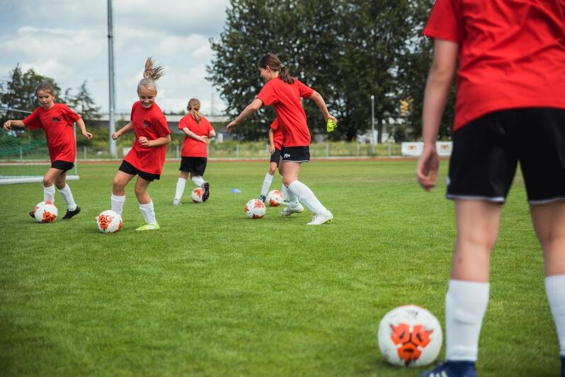 Młode kobiety na boisku w czerwonych koszulkach i czarnych spodenkach, jedna z nich ma przy nodze piłkę 