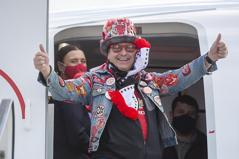 Uśmiechnięty mężczyzna wysiada z samolotu. Okulary na nosie, na głowie kolorowy (głównie czerwony) kapelusz, dżinsowa kurtka z emblematami, na szyi szalik w barwach Manchesteru, ręce do boku z uniesionymi kciukami  