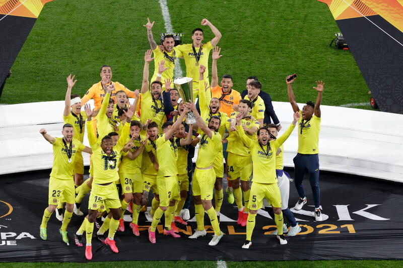 Piłkarze w żółtych strojach na stadionie w grupie. Cieszą się, skaczą, mają na szyjach medale, w środku dwóch trzyma w dłoniach do góry puchach 