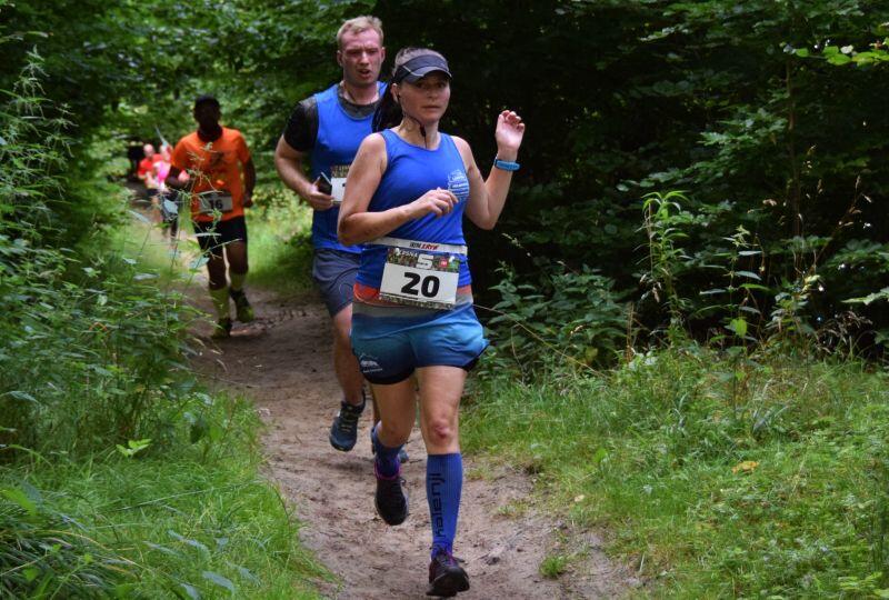 Kobieta ubrana w niebieski strój sportowy podczas biegu przez las, a nią biegną kolejni zawodnicy