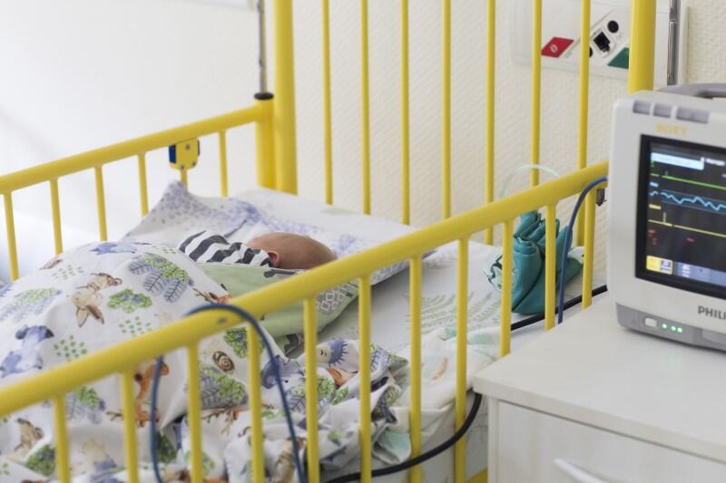 Sala szpitalna, łóżeczko dzięcięce, w środku śpi niemowlę przykryte kołdrą