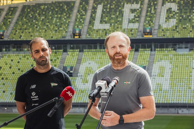 Dwaj mężczyźni stoją na murawie gdańskiego stadionu. Przed Piotrem Stokowcem znajdują się trzy statywy z mikrofonami. Flavio stoi na lewo od trenera
