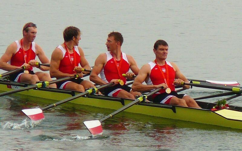 czwórka mężczyzn w sportowych strojach w łodzi z wiosłami w dłoniach, mają zawieszone na szyi złote medale