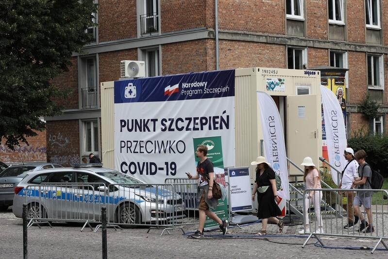 kremowy kontener z banerem z napisem narodowy punkt szczepień, obok policyjny radiowóz, kilka osób przechodzi w okolicy wejścia do konteneru
