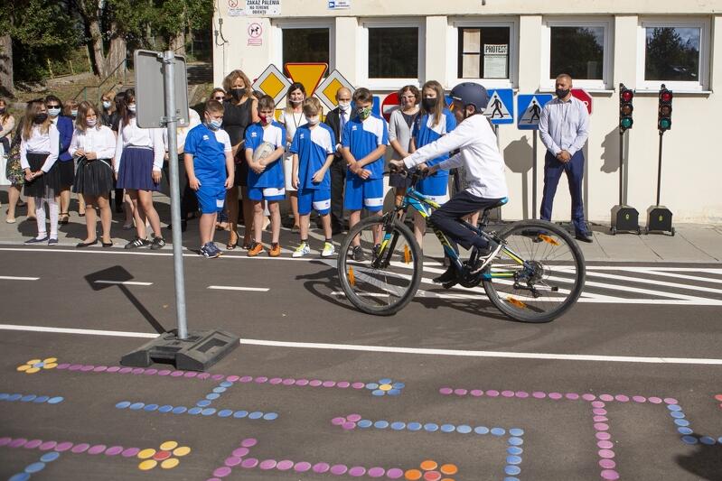 Grupa dzieci stojących w szeregu na boisku szkolnym. Przed nimi na rowerze przejeżdża uczeń w czarnym kasku na głowie, ubrany w białą koszulę i czarne spodnie. Rower skierowany jest w lewo