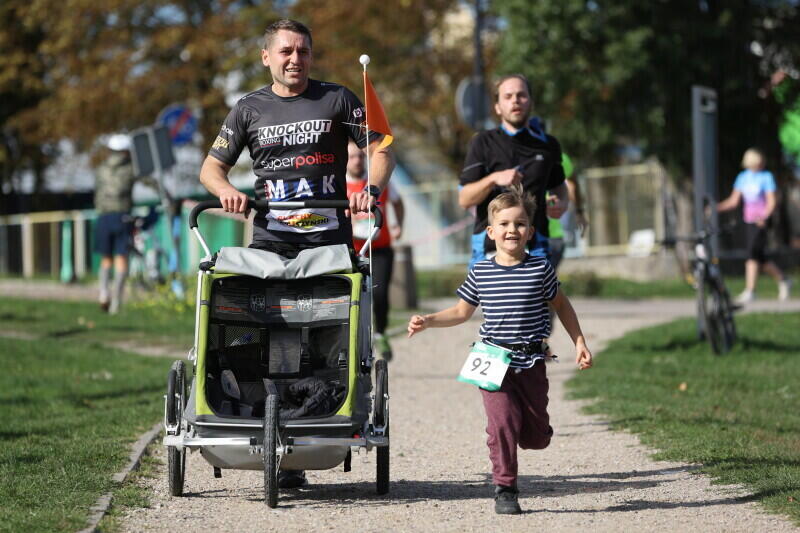 Biegnie mężczyzna w czarnej koszulce. Pcha przed sobą specjalny - przystosowany do biegania - wózek dla dziecka. Obok, po prawej stronie, biegnie około 5-letni chłopczyk z przypiętym numerem startowym 92
