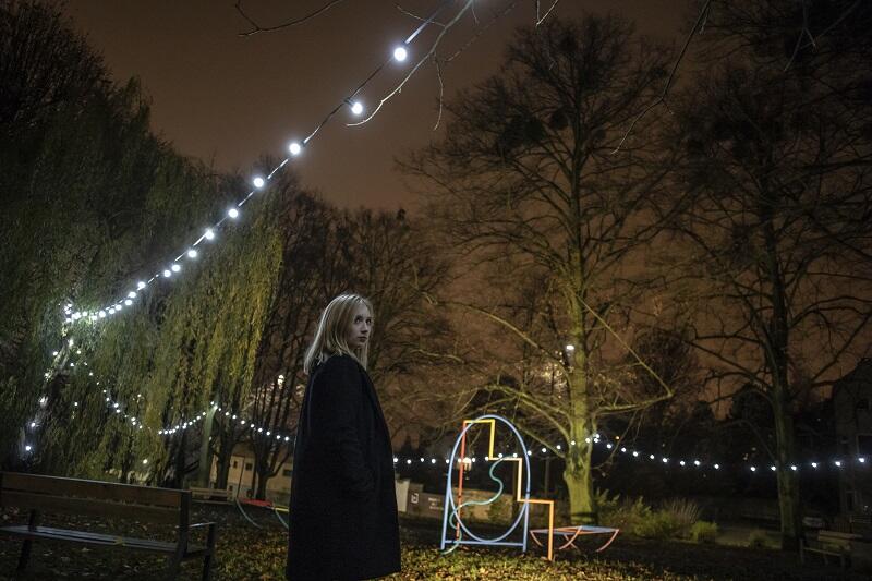 młoda kobieta blondynka ubrana w czarny płaszcz, stojąca w parku, na drzewach zwieszona jest girlanda z świecącymi lampkami