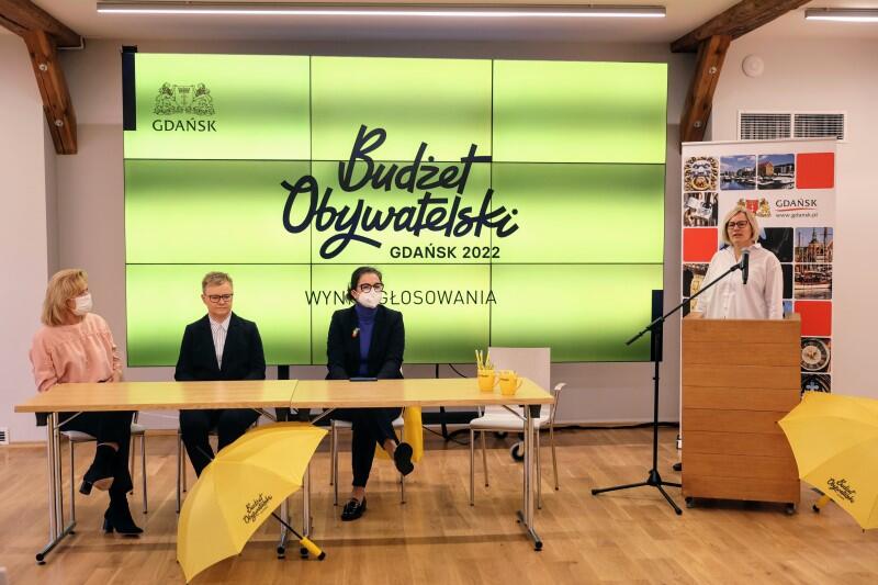 Cztery osoby dwie kobiety i mężczyzna siedzą przy stole, obok stoi kobiet przy mikrofonie w tle ekra z napisem Budziet Obywatelski Gdańsk 2022  