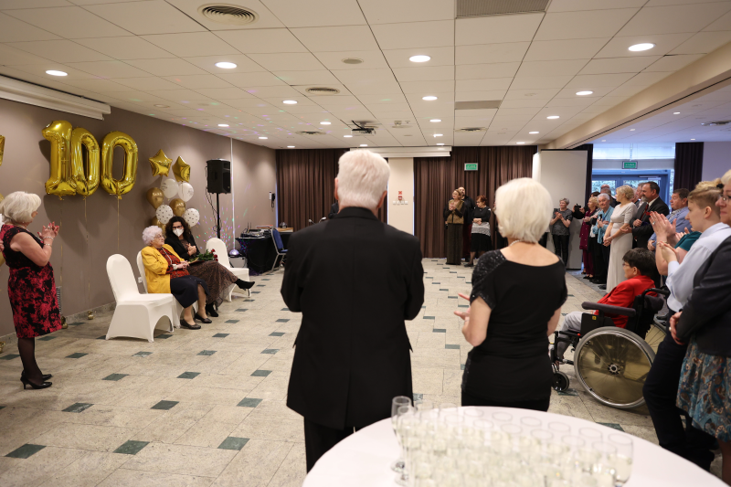 Sala hotelowa, część osób elegancko ubranych stoi, na fotelu siedzi starsza kobieta 
